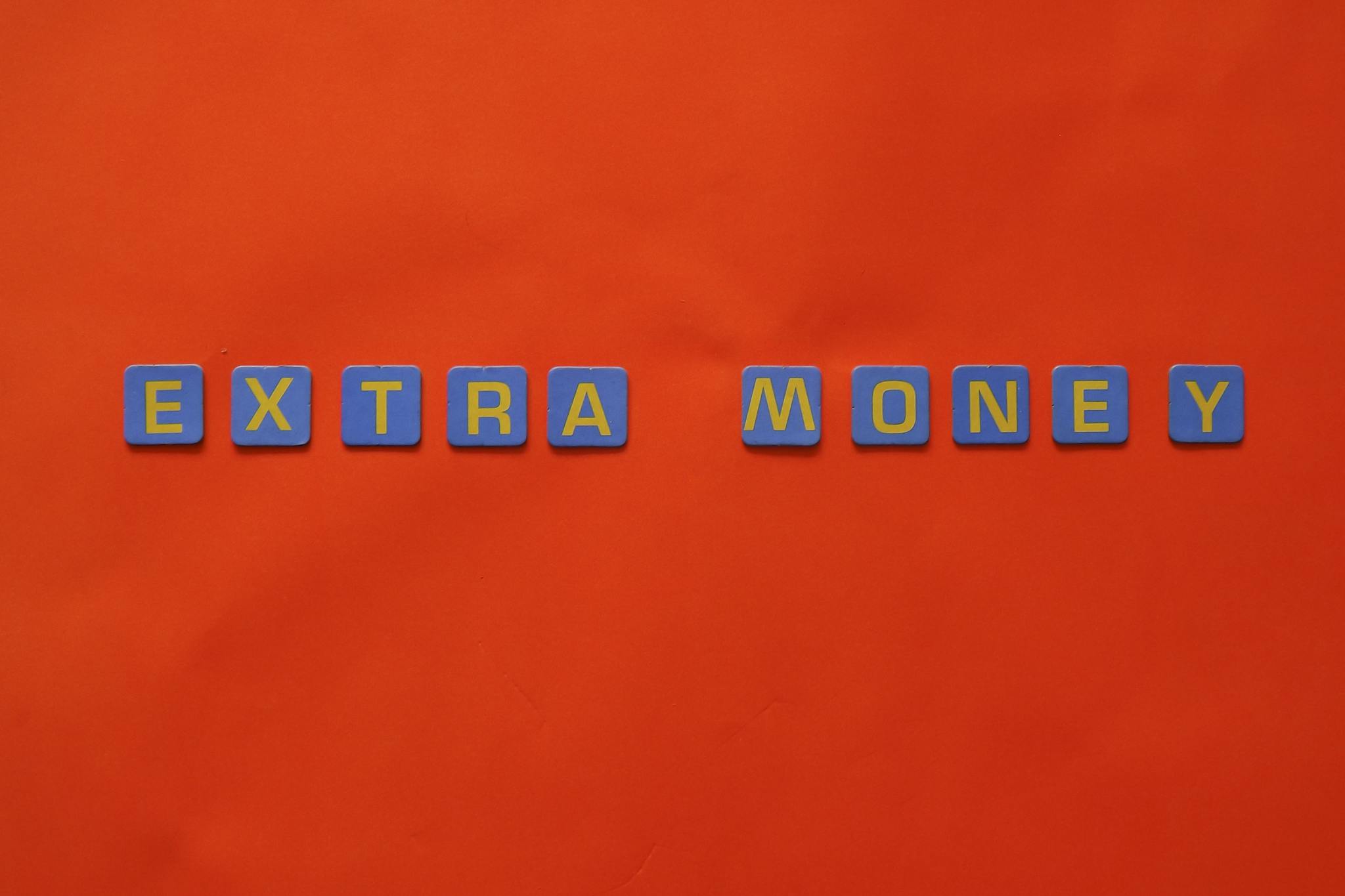 Bright orange background with 'EXTRA MONEY' spelled in blue and yellow tiles, illustrating financial concepts.